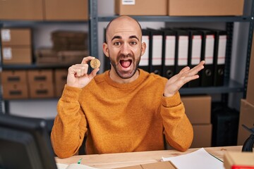 Canvas Print - Young bald man with beard working at small business ecommerce holding bitcoin celebrating achievement with happy smile and winner expression with raised hand