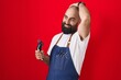 Leinwandbild Motiv Young hispanic man with beard and tattoos wearing barber apron holding razor smiling confident touching hair with hand up gesture, posing attractive and fashionable
