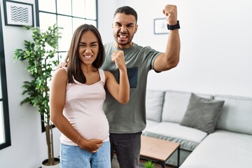 Canvas Print - Young interracial couple expecting a baby, touching pregnant belly angry and mad raising fist frustrated and furious while shouting with anger. rage and aggressive concept.