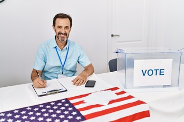 Wall Mural - Middle age man with beard at political election sitting by ballot looking positive and happy standing and smiling with a confident smile showing teeth