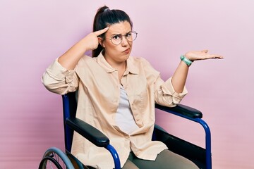 Poster - Young hispanic woman sitting on wheelchair confused and annoyed with open palm showing copy space and pointing finger to forehead. think about it.