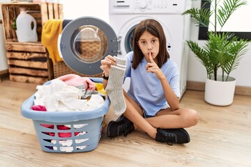 Sticker - Young hispanic girl doing laundry holding socks asking to be quiet with finger on lips. silence and secret concept.