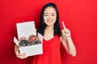 Leinwandbild Motiv Young chinese girl holding box with tasty colorful doughnuts smiling with an idea or question pointing finger with happy face, number one