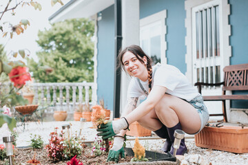 Cheerful young woman in gloves using gardening tools for planting flowers on back yard.casual clothes enjoying work at summer garden.Home garden new skills learning.Concept of gardening and floristics