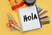 Notebook With Text HOLA (Spanish For HELLO), Flag Of Spain And Stationery On Yellow Background