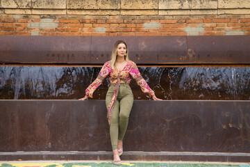 Pretty young blonde woman with sunglasses sitting on the edge of a fountain in the park. Holiday and leisure time concept.