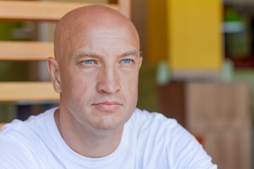 Portrait handsome bald man with blue eyes in a white t-shirt with a pensive look, closeup on the background in a cafe