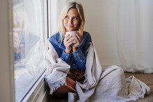 Young Woman Wrapped In Blanket Holding Coffee Cup Sitting By Window At Home
