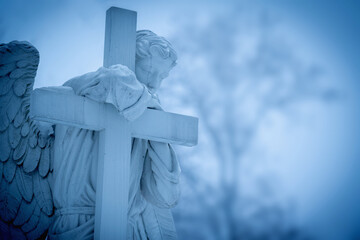 Fototapete - Old and ancient stone statue of angel with cross against blurred background of forest trees. Copy space.