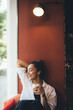 © polinaloves - Young woman drinking coffee cappuccino sitting near window.