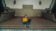© BublikHaus - Cinematic symmetric shot of young woman in yellow sweater sit on stairs of old university campus building, work on laptop on new exciting school project or assignment. Freshmen or junior student
