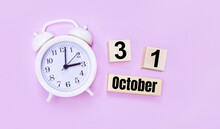 Two Wooden Cubes And A Block With The Text OCTOBER 31 And A White Alarm Clock On A Light Pink Background.