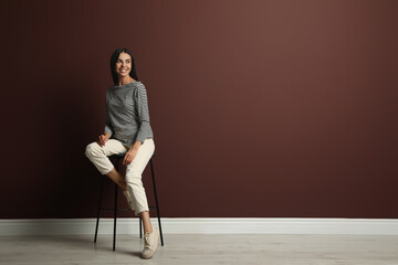 Beautiful young woman sitting on stool near brown wall. Space for text