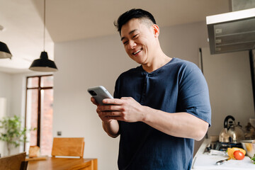 Wall Mural - Adult brunette asian man smiling and using cellphone at home