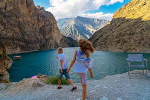 Back View Of Teen Boy And Girl Throwing Pebbles Into River Surrounded By Mountains At Summertime