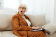 © Tatiana - a happy, pleasantly shocked elderly woman is sitting on a sofa in a bright interior and looks at the camera with her mouth wide open, holding a laptop on her lap