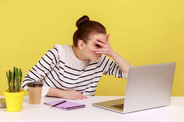 Curious woman manager sitting at workplace, spying, looking through fingers at laptop screen, peeking secret gossip in office. Indoor studio studio shot isolated on yellow background.