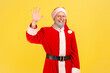 Positive good looking elderly man with gray beard wearing santa claus costume waving hand, greeting somebody, looking at camera with smile. Indoor studio shot isolated on yellow background.