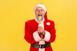 Excited elderly man with gray beard wearing santa claus costume holding and biting multicolored ice cream, celebrating winter holiday. Indoor studio shot isolated on yellow background.