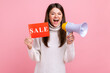 Excited brunette female holding sale card and megaphone, announcing discounts in shopping mall, wearing white casual style sweater. Indoor studio shot isolated on pink background.