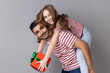 Portrait of extremely happy father and daughter in striped T-shirts celebrating holiday together, dad holding kid with present box on his back. Indoor studio shot isolated on gray background.