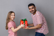 Portrait of optimistic smiling father and daughter in striped T-shirts looking at camera with happy expressing, dad giving birthday present to child. Indoor studio shot isolated on gray background.
