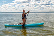 A shaved Jewish feminist woman in a closed swimsuit kneeling on a SUP board with an oar floats on the water.