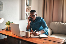 Young entrepreneur on his phone while taking notes at his desk. Professional man working remote from home with technology. African American male has a business meeting on an audio call.