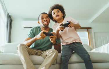 Happy Black African American Father and his Little son in Afro hair using joysticks or game controllers playing console video games together