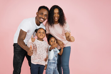 Poster - Happy family concept. Young parents and their daughters hugging sitting down on the floor showing thumbs up over pink studio background, free space.