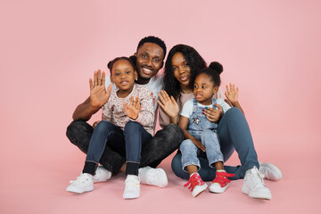 Poster - Portrait of smiling african american family of four sitting on floor waving their hands over pink studio background, free copy space, banner.