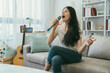 portrait of carefree asian girl sitting with folded legs is singing into microphone while having karaoke fun alone in a cozy living room at home with daylight.