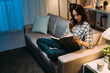 © PR Image Factory - beautiful chinese lady sitting comfortably on the couch is holding and reading a book alone on a peaceful night in a cozy home living room interior.