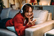 © PR Image Factory - carefree asian girl lying prone on sofa and propping her face is enjoying watching shows through streaming service on her phone in a comfortable home interior.