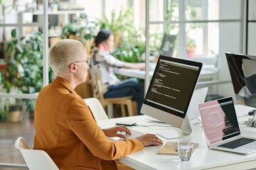 Rear view of female developer with short hair sitting at her workplace with computer monitor and typing codes