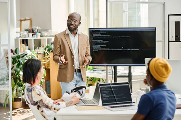 African programmer standing near the screen with codes and presenting new software for VR glasses to his colleagues at meeting