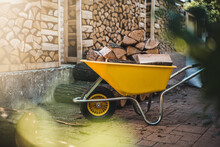 A Pile Of Split Firewood For Heating A House In The Yard, A Yellow Wheelbarrow With Firewood In The Foreground.