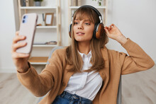 Joyful Young Blonde Lady In Warm Sweater In Headphones Listen Fav Songs Doing Selfie Video Call Sitting In Armchair At Modern Home Interior. Music Time Relaxing Cool Playlist Concept. Copy Space