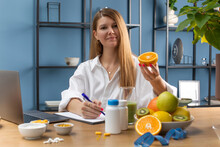  A Female Nutritionist Smiles At The Camera And Holds A Half Of An Orange, Sitting At Her Desk In A White Shirt.