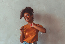 Happy African American Girl Standing With Festive Led Lights Around Hands And Body, Raising Up One Of Her Hand And Smiling On Camera, Posing Against Concrete Wall