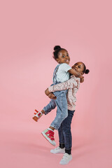 Wall Mural - Funny two little African cute girls sisters, having fun together, posing to camera one girl rises other in her arms, on isolated pink color studio background.