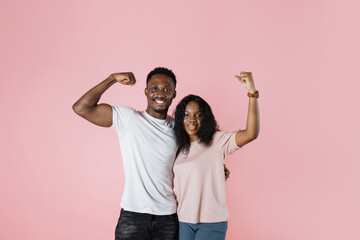 Wall Mural - Young cheerful strong sporty fitness african couple two friends man woman in casual t-shirts showing biceps muscles on hand isolated on pastel pink color background studio portrait.