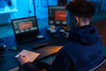 cybercrime, hacking and technology concept - close up of male hacker in dark room writing to noteboo
