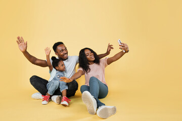 Family fun. Young african american parents spending time with their little daughter with open arms using phone for selfie over yellow background, banner, panorama, copy space.