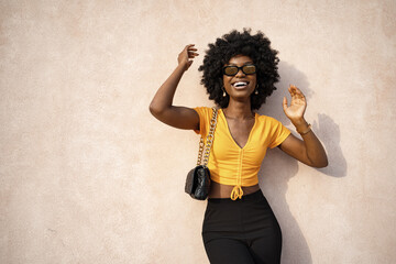 Happy African American young woman with afro hair wearing stylish sunglasses posing against beige wall background