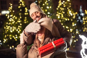 Wall Mural - winter holidays and people concept - happy smiling couple with gift over christmas tree lights in evening city