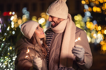 Poster - winter holidays and people concept - happy smiling couple with sparklers over christmas lights in evening