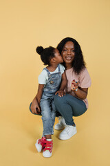 Wall Mural - Motherhood and family concept. Lovely cute little African girl kid in summer outfits, having fun and kissing her happy smiling young pretty mother, posing on isolated yellow background