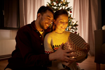 Poster - holidays, celebration and people concept - happy couple opening christmas gift box with magical light inside at home