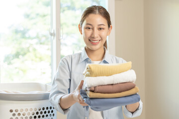 Wall Mural - Housewife, attractive asian young woman, girl holding many folding clothes in the basket after washing clean clothes and drying, Household working in room at home, maid chore laundry tidy.